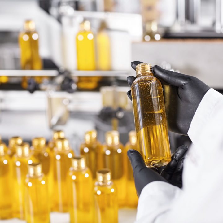 Horizontal portrait of mass process at factory. Researcher standing near many bottles with yellow liquid holding one of them isolated over factory conveyor. Selective focus. Great success of factory